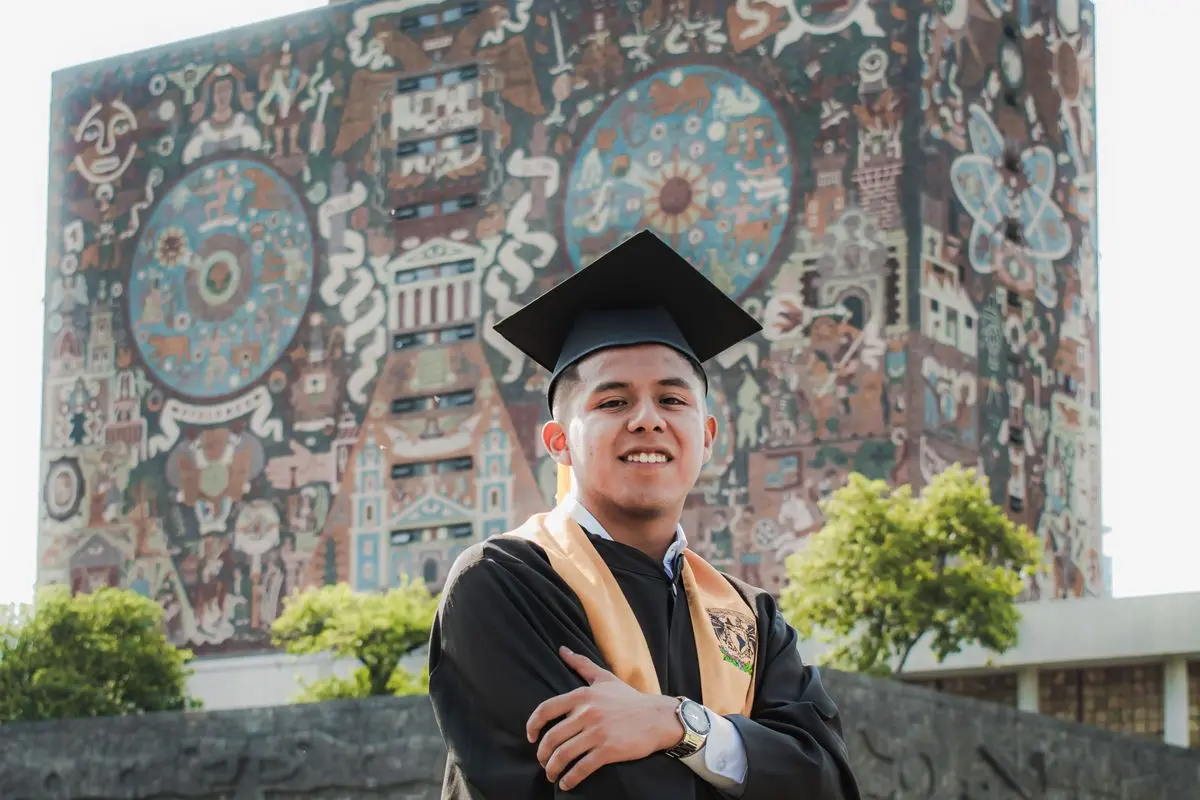 Egresado celebrando su graduación con birrete y toga frente al mural de la UNAM